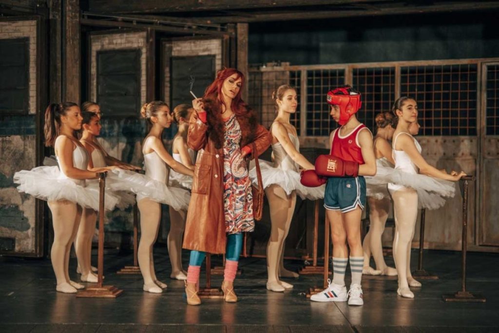 Donna coi capelli rossi  e dietro giovani ballerini e un ragazzo vestito da pugile