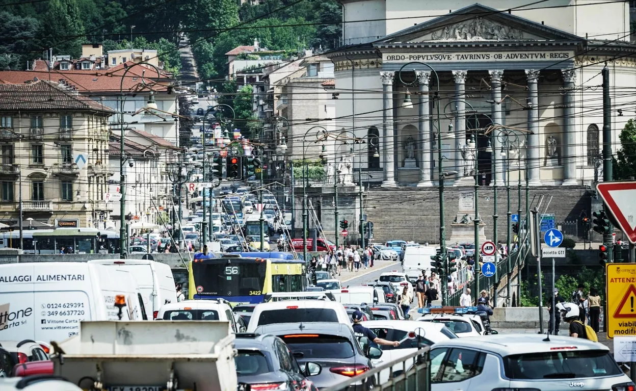 raffico intenso a Torino sul Ponte Vittorio Emanuele I, con auto, autobus e tram bloccati nel congestionamento. Sullo sfondo si vede la Chiesa della Gran Madre di Dio, con numerosi pedoni che attraversano la strada. L'immagine mostra una scena urbana caratterizzata da cavi elettrici del trasporto pubblico e segnaletica stradale