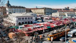 Vista aerea del mercato di Porta Palazzo a Torino, con le sue bancarelle coperte da tende colorate e la Tettoia dell'Orologio restaurata, circondata da edifici storici e mezzi pubblici
