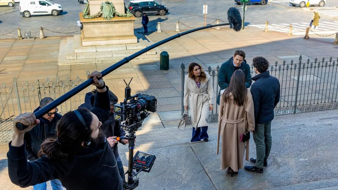Riprese di una scena cinematografica all'aperto con attori impegnati in un dialogo su una scalinata. Un operatore regge un'asta con microfono direzionale mentre una telecamera professionale registra la scena