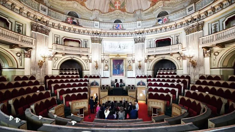 Interno della Camera dei Deputati del Parlamento Subalpino a Palazzo Carignano, Torino, con sedute in velluto rosso, decorazioni dorate e il ritratto di Vittorio Emanuele II al centro. Un gruppo di visitatori esplora l’aula storica