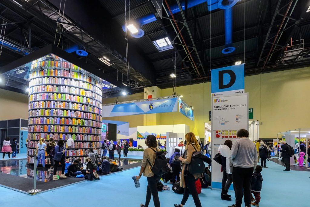 Visitatori al Padiglione 1 del Salone del Libro di Torino 2025, con la celebre torre di libri e lo stand della Città di Torino sullo sfondo.