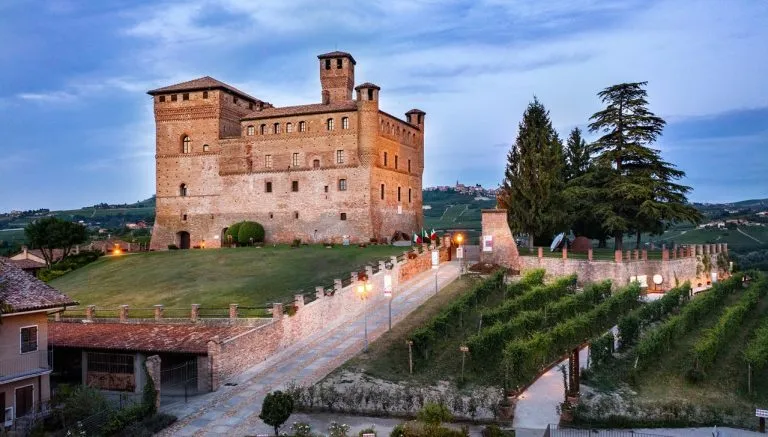 Veduta serale del castello di Grinzane Cavour con luci accese, filari di vigna in primo piano e colline delle Langhe sullo sfondo