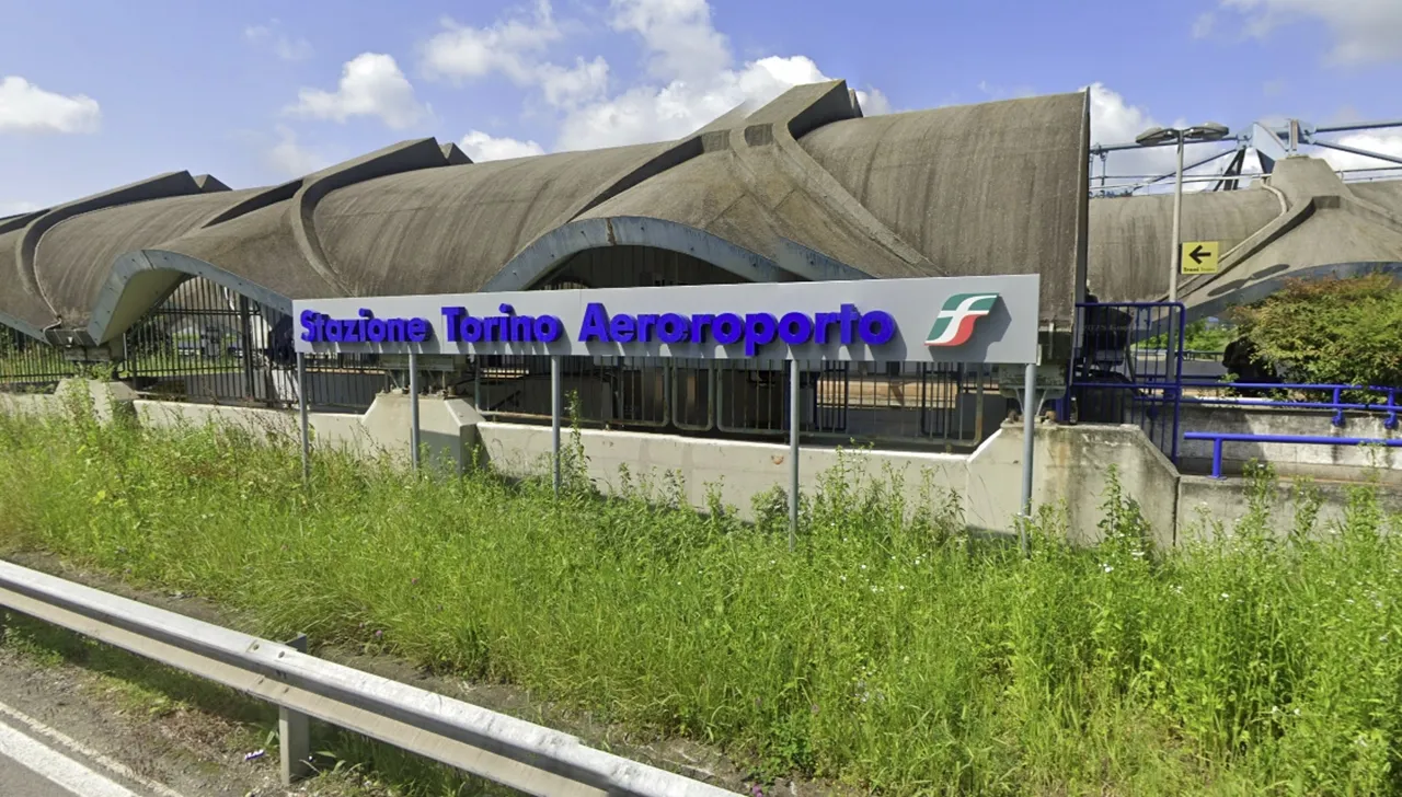 Facciata della stazione ferroviaria Torino Aeroporto, con l’insegna “Stazione Torino Aeroporto” in caratteri blu e logo di Trenitalia, davanti a una struttura moderna con archi in cemento. Vegetazione spontanea in primo piano.