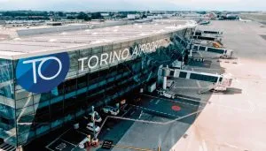 Vista panoramica dell’aeroporto di Torino Caselle (Sandro Pertini) con finger e jet bridge collegati al terminal, evidenziato il logo TO Torino Airport su facciata vetrata