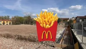 Area di cantiere in corso Potenza a Torino dove sorgerà un nuovo ristorante McDonald’s: il terreno è stato livellato e recintato, con lavori in corso lungo un canale di scavo. In primo piano è stata sovrapposta digitalmente un’icona di patatine fritte McDonald’s. Sullo sfondo si intravedono edifici residenziali e alberi sotto un cielo parzialmente nuvoloso
