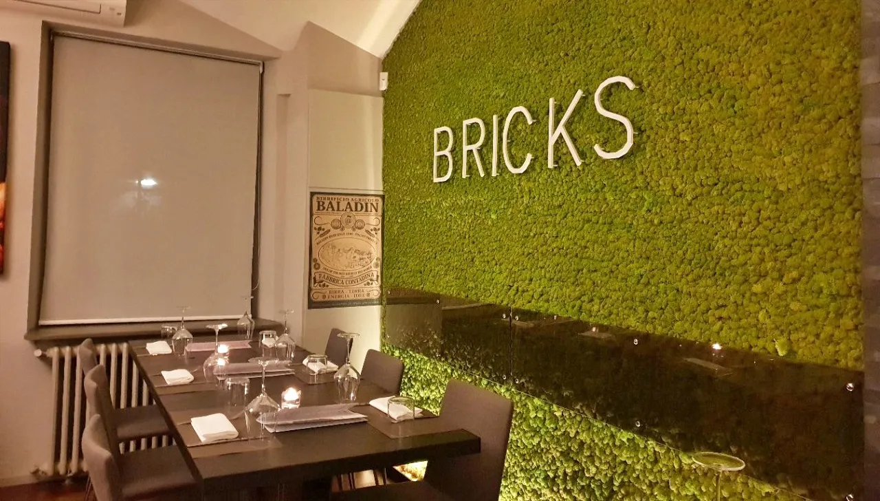 Tavoli apparecchiati all’interno della pizzeria Bricks di Torino, con parete verde verticale decorativa e insegna luminosa con la scritta