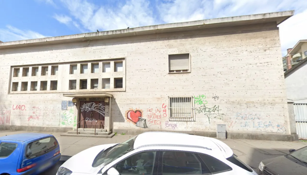 Facciata esterna dei bagni municipali abbandonati di via Roccavione a Torino, edificio in muratura chiara con scritte vandaliche, ingresso sbarrato e finestra con inferriata, auto parcheggiate lungo la strada
