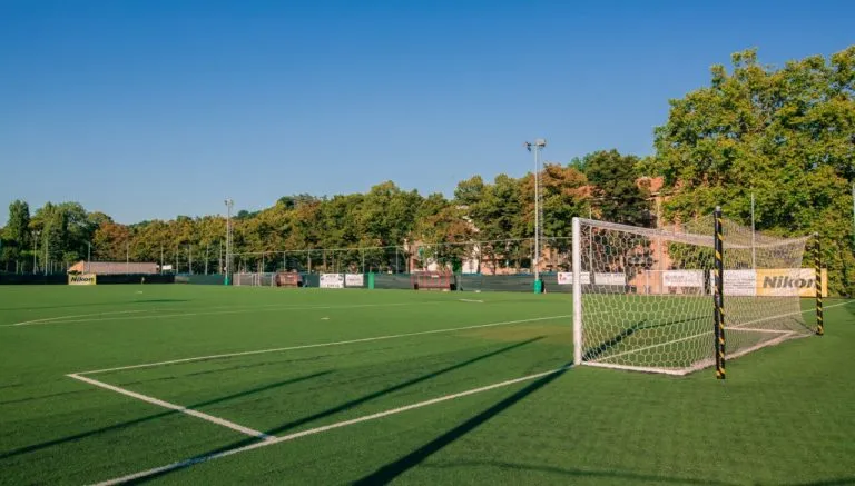Campo da calcio in erba sintetica della CBS Torino, con rete da porta in primo piano e tribune sullo sfondo, immerso nel verde di corso Sicilia.