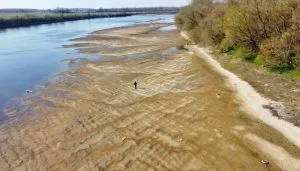 Vista aerea di un tratto del fiume Po con portata ridotta e ampie zone di letto asciutto, segno di siccità, con una persona che cammina sul terreno sabbioso e alberi lungo la riva.