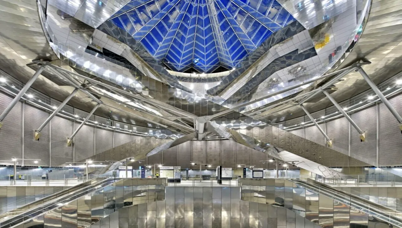 Interno futuristico di una stazione metropolitana con struttura architettonica in acciaio e vetro, cupola trasparente con travi incrociate e illuminazione moderna