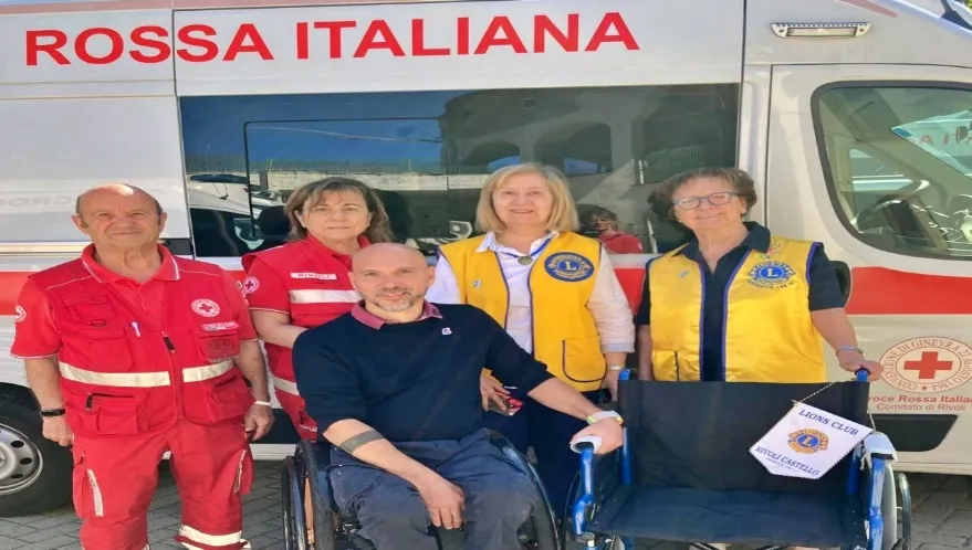 Volontari della Croce Rossa Italiana e membri del Lions Club posano insieme davanti a un’ambulanza, accanto a una sedia a rotelle con bandiera Lions