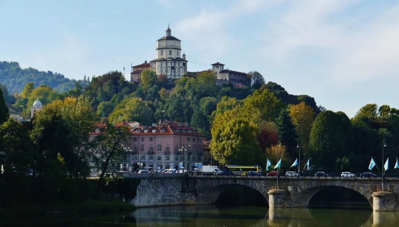 Veduta panoramica del Monte dei Cappuccini a Torino, con la chiesa barocca in cima alla collina circondata da alberi verdeggianti. In primo piano, il ponte Vittorio Emanuele I attraversa il fiume Po, con traffico urbano e bandiere azzurre esposte lungo il parapetto. Sullo sfondo, cielo sereno e atmosfera autunnale