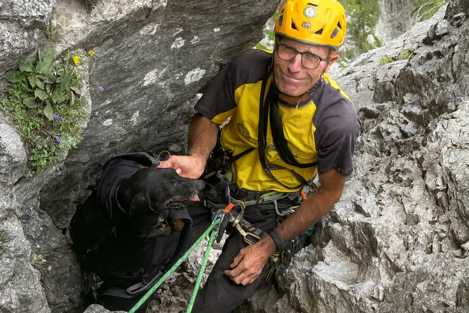 Un soccorritore alpino, con casco giallo e imbragatura, recupera un cane nero intrappolato in un canalone roccioso a Bardonecchia durante un’operazione di salvataggio