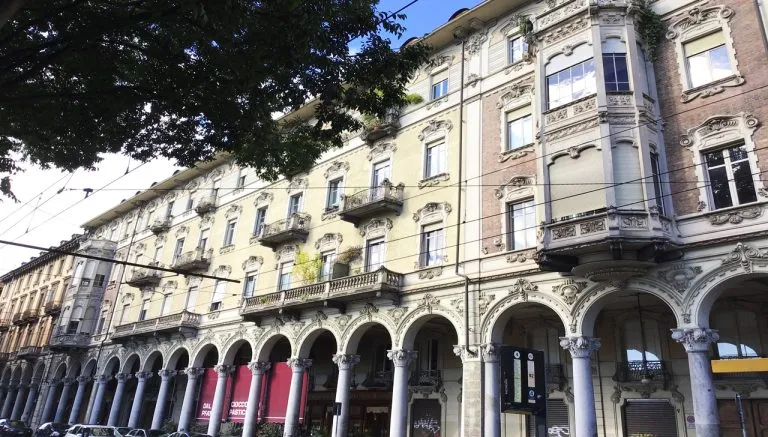 Facciata liberty di via Sacchi a Torino, con portici ad archi, colonne in pietra e decorazioni elaborate su balconi e finestre, sotto una linea di alberi e fili del tram