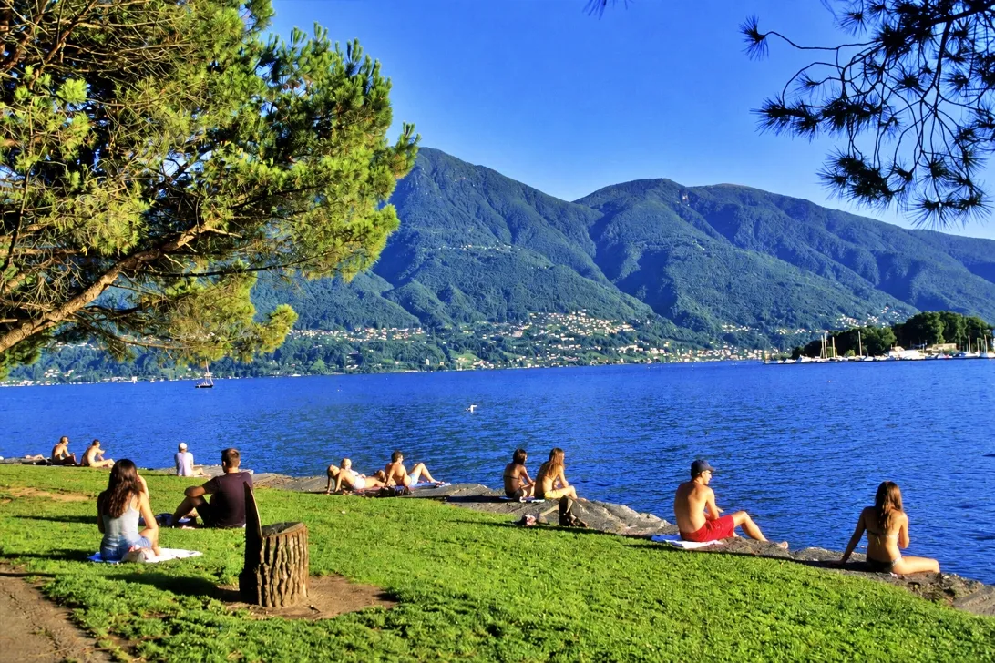 Persone rilassate sulla riva del lago, tra prato e alberi, con le montagne verdi e i paesi affacciati sull’acqua sullo sfondo.